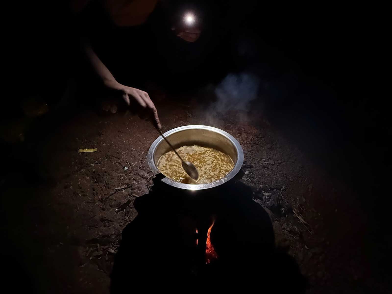 A person stirs a steaming pot of Maggi noodles cooking over a small, glowing fire on the ground, illuminated by a headlamp, during a pre-dawn snack session at the Savlya Ghat campsite.